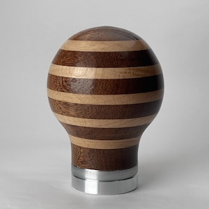 May include: A decorative wooden sphere featuring alternating bands of light and dark brown wood. The sphere is mounted on a silver metal base. The wood grain is visible, enhancing the handcrafted aesthetic. This item could be used as a unique home decor accent.