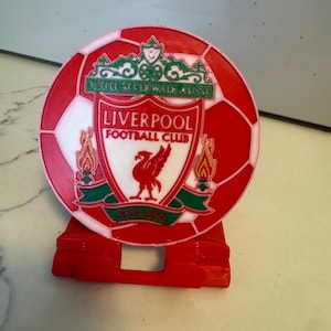 May include: A 3D printed football with the Liverpool Football Club logo. The logo features a red bird, a white shield, and the text "Liverpool Football Club" and "Est 1892". The ball is on a red stand.