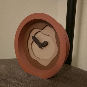 May include: A round, terracotta-colored clock with a layered, organic design. The clock face features concentric rings of varying brown and beige tones. The hands are dark brown, indicating the time. The clock is a decorative home decor item.