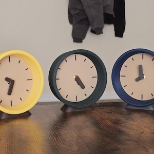 May include: Three modern analog clocks in yellow, teal, and blue. Each clock has a light beige face with black hour markers and hands. The clocks are on a dark brown wooden surface.