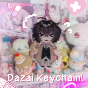 May include: A clear acrylic Dazai keychain featuring a cartoon character with brown hair, wearing a tan coat and black outfit. The keychain has a pink noose design and the text "Dazai Keychain!" in white. The background is a soft pink.