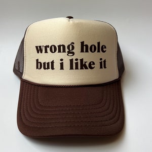 May include: A brown and beige trucker hat with the text "wrong hole but i like it" in brown lettering.
