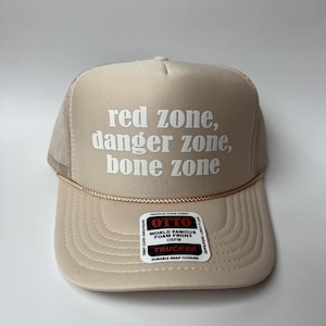 May include: A beige trucker cap featuring the text "red zone, danger zone, bone zone" in white. The hat has a mesh back and a curved peak. The label reads "OTTO" and "TRUCKER".
