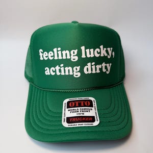 Peut inclure: Casquette de camionneur verte avec l'inscription blanche "feeling lucky, acting dirty". Le chapeau a un dos en filet et une visière incurvée. Une étiquette blanche indique "OTTO WORLD FAMOUS FOAM FRONT TRUCKER".