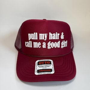 May include: Burgundy trucker hat with white text that reads "pull my hair & call me a good girl." The hat has a mesh back and a snap closure. A white sticker on the front reads "OTTO WORLD FAMOUS FOAM FRONT OSFM TRUCKER DURABLE SNAP CLOSURE."