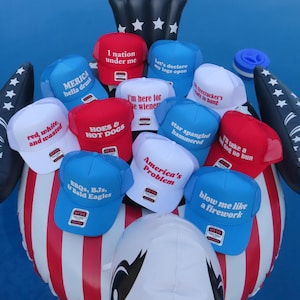 May include: A selection of red, white, and blue trucker hats with various slogans, such as "America's Problem" and "Blow me like a firework." The hats are arranged on an inflatable eagle, evoking a patriotic theme.