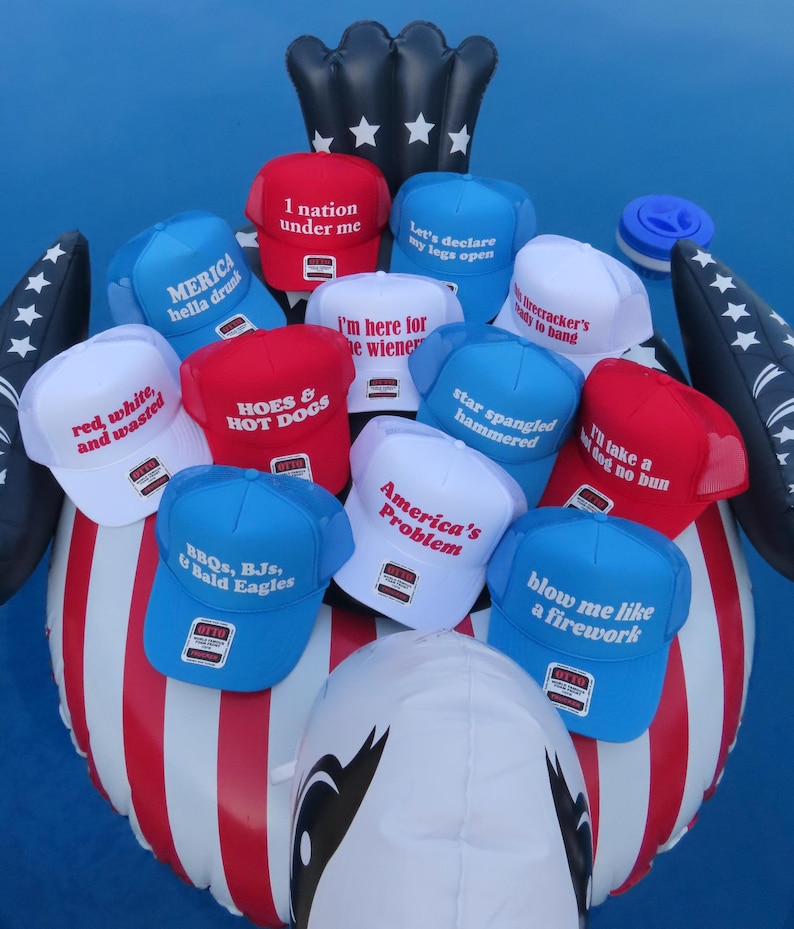 May include: A selection of red, white, and blue trucker hats with various slogans, such as "America's Problem" and "Blow me like a firework." The hats are arranged on an inflatable eagle, evoking a patriotic theme.