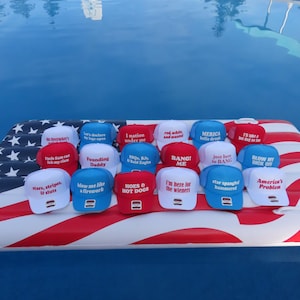 May include: A selection of baseball caps in red, white, and blue, each bearing a different slogan. The caps are displayed on an inflatable raft decorated with the American flag, with a swimming pool in the background. The caps feature phrases like "America's Problem" and "I'm here for the wieners."