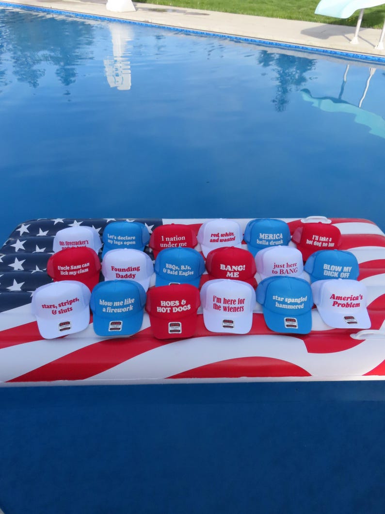 May include: A selection of baseball caps in red, white, and blue, each bearing a different slogan. The caps are displayed on an inflatable raft decorated with the American flag, with a swimming pool in the background. The caps feature phrases like "America's Problem" and "I'm here for the wieners."