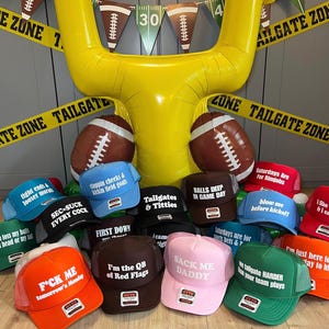 May include: A collection of multi-colored trucker hats featuring various humorous text prints, arranged in front of a large yellow inflatable goal post and football balloons, with yellow caution tape banners reading TAILGATE ZONE.