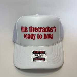 May include: White trucker hat with the text "this firecracker's ready to bang" in red. The hat features a white foam front and a mesh back. The hat has a snap closure and a terry cloth sweatband.