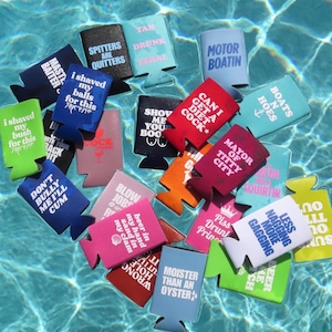 Könnte beinhalten: Eine Sammlung bunter Getränkedosenkühler, die im Wasser schwimmen. Jeder Kühler hat ein einzigartiges Design mit Text, darunter Sätze wie "Motor Boatin" und "Moister Than An Oyster". Die Kühler sind in verschiedenen Farben wie Blau, Rosa und Grün.