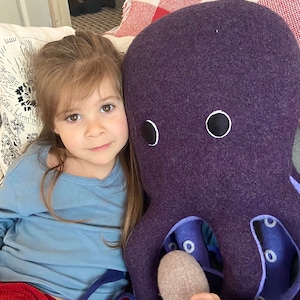 May include: A young girl is cuddling a large purple plush octopus with black eyes. The octopus has a light purple underside with white circles.