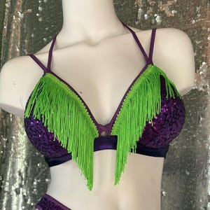 May include: A purple sequin bikini set, comprising a halter-neck top with lime green fringe and high-cut bottoms. The set is displayed on a white mannequin. The background is a silver sequin fabric.