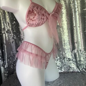 May include: A pink velvet and tulle lingerie set with a bow detail. The top is a triangle bra with adjustable straps and the bottom is a high-waisted panty with a ruffled trim.