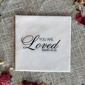 May include: A square white napkin featuring black printed text that reads YOU ARE Loved and ISAIAH 41:10. The word Loved is written in a large, elegant cursive script, while the other words are in a clean, simple sans-serif font.