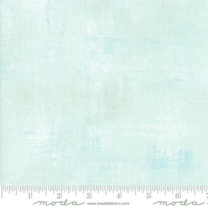 May include: A light blue and white fabric with a textured, painterly effect. The fabric has a subtle, mottled appearance with a ruler along the bottom edge. The word "moda" and website address are printed at the bottom.