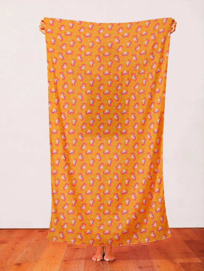 May include: A yellow fabric with a repeating pattern of pink and purple unicorn heads. The fabric is being held up by a person's hands.