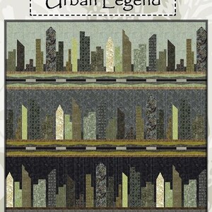 May include: A fabric quilt with a cityscape design. The quilt features a repeating pattern of stylized buildings in shades of green, brown, and beige. The text "Urban Legend" is at the top of the quilt.