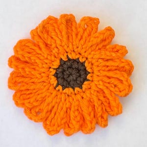 May include: A crocheted orange flower with a brown center. The flower is made with yarn and has a textured appearance.