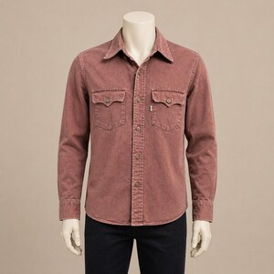 Puede incluir: Camisa vaquera de color rosa polvoriento de manga larga, con dos bolsillos en el pecho y cierre de botones. La camisa tiene un cuello clásico y se muestra en un maniquí. La camisa se combina con unos vaqueros azul oscuro.