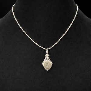 May include: Silver-toned pendant necklace with a delicate chain. The pendant features an ornate design with a circular center, displaying the letter "e". The necklace is displayed against a black background.