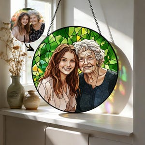 May include: A round stained glass art piece featuring a portrait of two smiling women. The background is a mosaic of green glass. The artwork is suspended by a black chain. A smaller photo of the women is in the upper left corner.