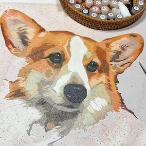 May include: A paint-by-numbers kit featuring a close-up of a Corgi dog's face. The dog has a white and brown coat with a black nose and eyes. A wicker basket with numbered paint pots and a paintbrush are in the background.