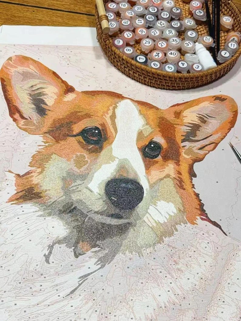May include: A paint-by-numbers kit featuring a close-up of a Corgi dog's face. The dog has a white and brown coat with a black nose and eyes. A wicker basket with numbered paint pots and a paintbrush are in the background.