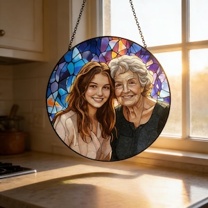 May include: Round stained glass window art featuring two smiling women. The artwork has a black metal frame and a chain for hanging. The background is a colorful mosaic of blue, purple, and orange glass. The women are depicted in a realistic style.