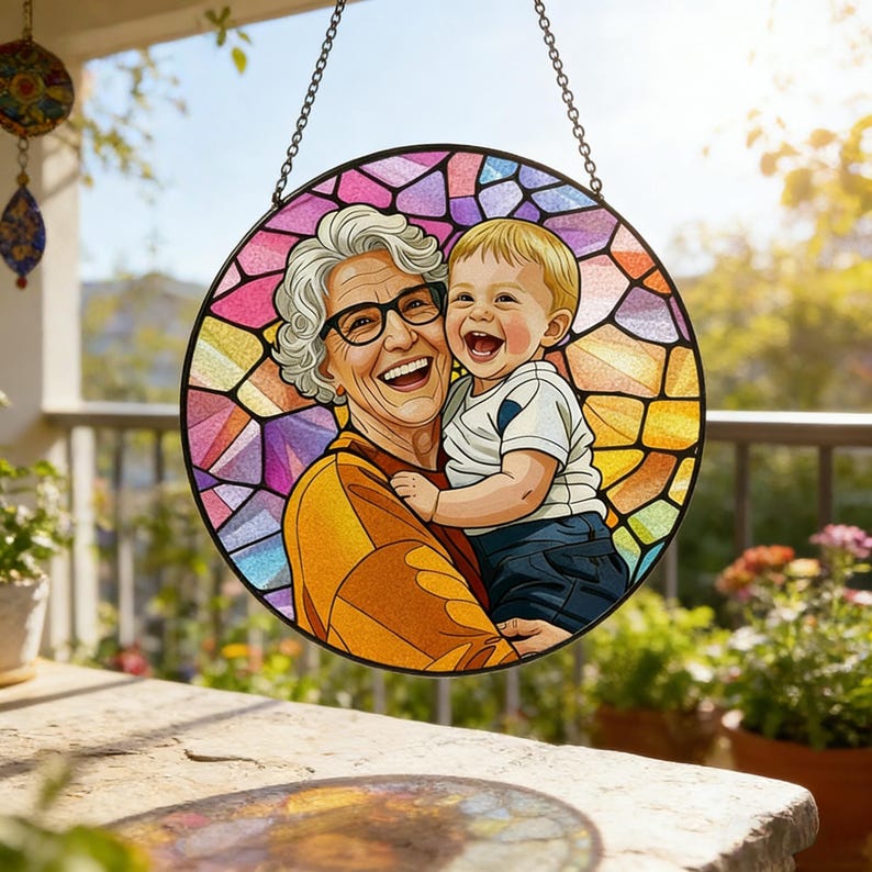 May include: A round stained glass window featuring a smiling woman wearing glasses holding a laughing baby. The background is a colorful mosaic pattern. The window is hanging from a chain.