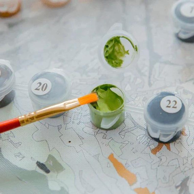 May include: A paint-by-numbers kit with small paint pots and a paintbrush. The paintbrush has an orange tip and a red handle. The paint pots are labeled with numbers, and one pot contains green paint. The kit is on a patterned surface.