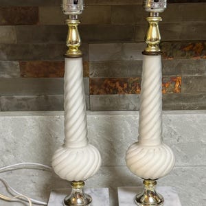 May include: Pair of vintage table lamps. Each lamp features a white, swirled glass column, a gold-colored metal accent, and a square marble base. The lamps are designed for use with light bulbs.