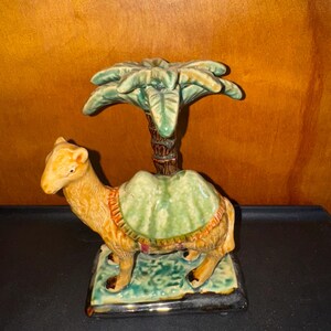 May include: A ceramic camel figurine with a green and brown palm tree on its back. The camel is standing on a green and brown base.