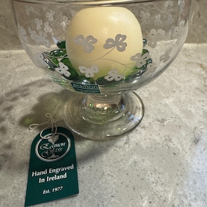 May include: A clear glass bowl with a pedestal base, etched with a shamrock design. Inside, a large cream-colored candle rests on green glass beads. A tag reads "Eamon Glass, Hand Engraved In Ireland, Est. 1977."