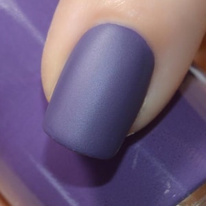 May include: A close-up of a purple nail polish bottle with a matte finish. The bottle is held by a person, with one nail painted to match the polish. The nail polish has a matte finish and a deep purple color.