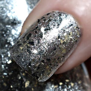 May include: Close-up of a fingernail painted with silver glitter nail polish. The polish has a mix of silver, black, and gold glitter flakes. The nail is held over a bottle of the same nail polish.
