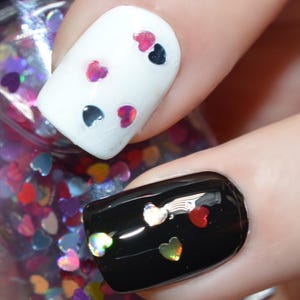May include: Close-up of a nail polish bottle and manicured nails. One nail is painted white with heart-shaped glitter, and another is black with heart-shaped glitter. The nail polish bottle contains heart-shaped glitter.
