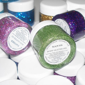May include: Close-up of several small jars of body glitter gel. The jars are labeled with the names "Panther", "Mantis", and "Magic". The glitter is in various colors, including pink, green, gold, and purple.