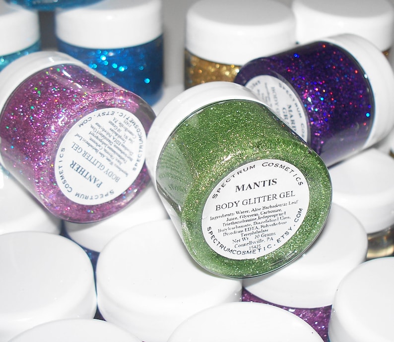 May include: Close-up of several small jars of body glitter gel. The jars are labeled with the names "Panther", "Mantis", and "Magic". The glitter is in various colors, including pink, green, gold, and purple.