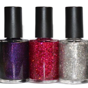 May include: Three nail polish bottles with black caps. The bottles contain purple, red, and silver glitter polish. The bottles are clear, allowing the color of the polish to be seen. The background is white.