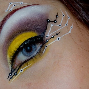 May include: Close-up of a woman's eye with yellow, black, and white makeup. The makeup design features a geometric pattern with white lines and black dots.