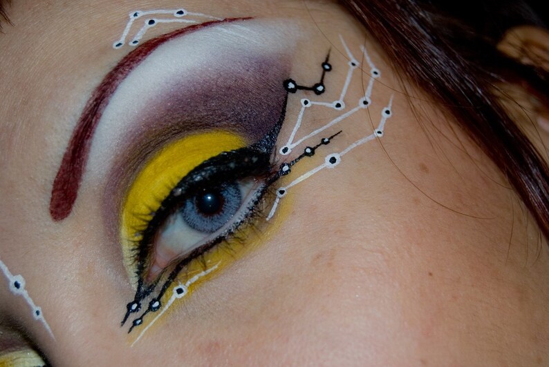 May include: Close-up of a woman's eye with yellow, black, and white makeup. The makeup design features a geometric pattern with white lines and black dots.