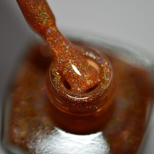 May include: A close-up of a bottle of nail polish with a gold glitter finish. The bottle is open and the brush is dipped in the polish.