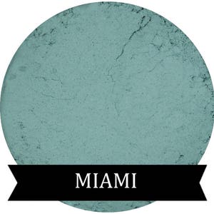 May include: A round, teal-coloured eyeshadow with a textured surface. The text "MIAMI" is printed in white on a black banner below the eyeshadow.