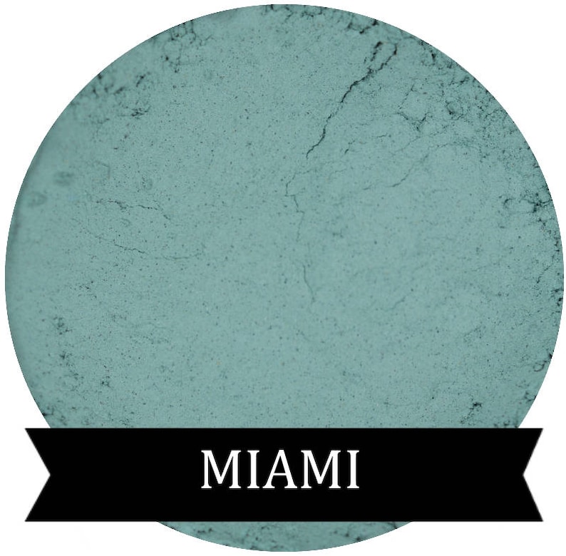 May include: A round, teal-coloured eyeshadow with a textured surface. The text "MIAMI" is printed in white on a black banner below the eyeshadow.