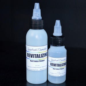 May include: Two bottles of Spectrum Cosmetics Revitalizing Nail Polish Thinner. The bottles are light blue with black caps and white labels. The labels read "Revitalizing Nail Polish Thinner" and list ingredients. The larger bottle is 2 fl oz.