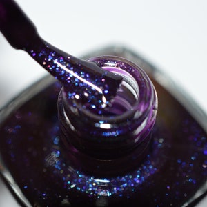 May include: A close-up of a bottle of dark purple nail polish with a brush dipped in the polish. The polish has a glittery finish.