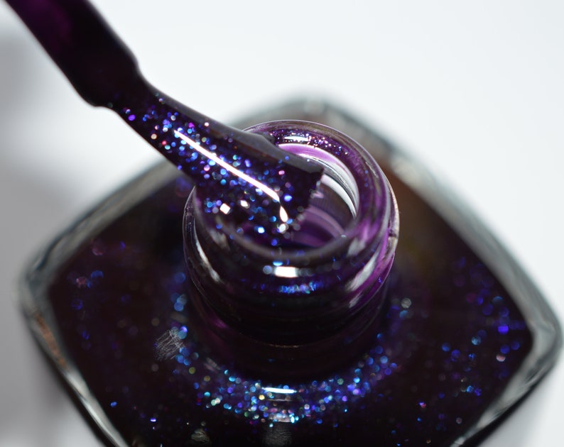 May include: A close-up of a bottle of dark purple nail polish with a brush dipped in the polish. The polish has a glittery finish.