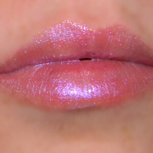 May include: Close-up of pink lips with a clear, iridescent lip gloss. The gloss has a subtle shimmer and reflects light.